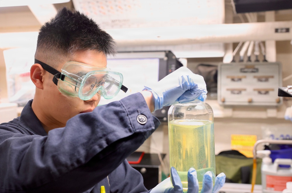 A young male laboratory technician or scientist is closely examining a large glass container filled with a yellow-green liquid. He is wearing appropriate personal protective equipment including safety goggles and blue nitrile gloves. The setting appears to be a professional or institutional laboratory environment.