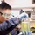 A young male laboratory technician or scientist is closely examining a large glass container filled with a yellow-green liquid. He is wearing appropriate personal protective equipment including safety goggles and blue nitrile gloves. The setting appears to be a professional or institutional laboratory environment.