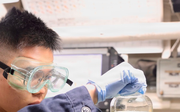 A young male laboratory technician or scientist is closely examining a large glass container filled with a yellow-green liquid. He is wearing appropriate personal protective equipment including safety goggles and blue nitrile gloves. The setting appears to be a professional or institutional laboratory environment.