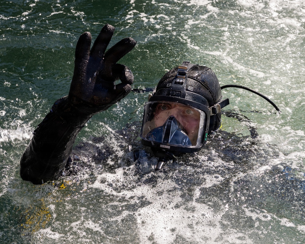 A diver is partially submerged in choppy, foamy water while giving an 'OK' hand signal with their right hand. The individual is wearing full tactical/professional diving gear including a full-face diving mask, black neoprene wetsuit, black diving gloves, and a helmet with mounted equipment. The image is sharp and well-lit, capturing water droplets and surface turbulence in detail.