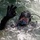 Forged Underwater: PNSY Dive Team Conducts Inspection of New Caisson A diver is partially submerged in choppy, foamy water while giving an 'OK' hand signal with their right hand. The individual is wearing full tactical/professional diving gear including a full-face diving mask, black neoprene wetsuit, black diving gloves, and a helmet with mounted equipment. The image is sharp and well-lit, capturing water droplets and surface turbulence in detail.