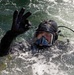 A diver is partially submerged in choppy, foamy water while giving an 'OK' hand signal with their right hand. The individual is wearing full tactical/professional diving gear including a full-face diving mask, black neoprene wetsuit, black diving gloves, and a helmet with mounted equipment. The image is sharp and well-lit, capturing water droplets and surface turbulence in detail.
