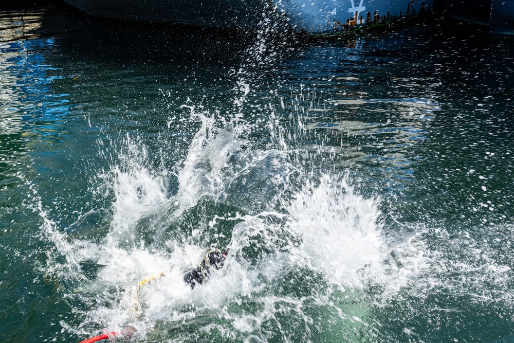 The image captures a dramatic water splash event, likely a person or object entering a body of water at high speed near a large vessel or dock structure. The foreground shows an explosive white water splash with what appears to be a person in dark clothing partially submerged. The background reveals calm green-blue water and the dark hull of a large ship or dock infrastructure.