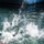 Forged Underwater: PNSY Dive Team Conducts Inspection of New Caisson The image captures a dramatic water splash event, likely a person or object entering a body of water at high speed near a large vessel or dock structure. The foreground shows an explosive white water splash with what appears to be a person in dark clothing partially submerged. The background reveals calm green-blue water and the dark hull of a large ship or dock infrastructure.
