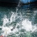 The image captures a dramatic water splash event, likely a person or object entering a body of water at high speed near a large vessel or dock structure. The foreground shows an explosive white water splash with what appears to be a person in dark clothing partially submerged. The background reveals calm green-blue water and the dark hull of a large ship or dock infrastructure.