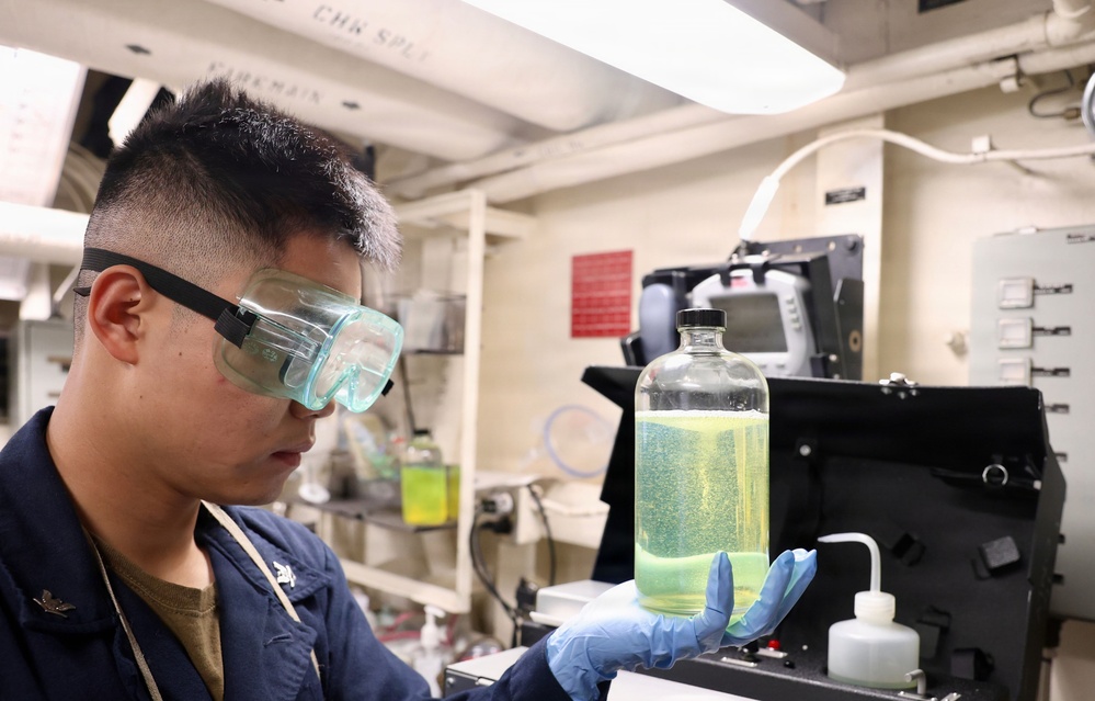 A person wearing safety goggles and blue nitrile gloves is examining a glass bottle containing a yellow-green liquid in what appears to be a laboratory or technical workspace. The individual is dressed in a dark blue uniform, suggesting a military or institutional affiliation. The setting appears to be an onboard ship laboratory based on the low ceiling, pipes, and compact equipment arrangement.