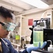 A person wearing safety goggles and blue nitrile gloves is examining a glass bottle containing a yellow-green liquid in what appears to be a laboratory or technical workspace. The individual is dressed in a dark blue uniform, suggesting a military or institutional affiliation. The setting appears to be an onboard ship laboratory based on the low ceiling, pipes, and compact equipment arrangement.