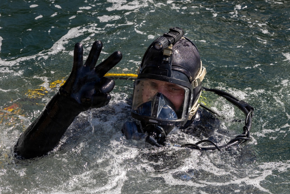 A diver is partially surfaced in open water, wearing full black tactical diving gear including a full-face mask and helmet-mounted light. The diver is making a hand signal with three fingers raised, likely communicating status or a number to someone off-camera. The image is sharp and well-lit, capturing fine details of the equipment and water surface.