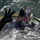Forged Underwater: PNSY Dive Team Conducts Inspection of New Caisson A diver is partially surfaced in open water, wearing full black tactical diving gear including a full-face mask and helmet-mounted light. The diver is making a hand signal with three fingers raised, likely communicating status or a number to someone off-camera. The image is sharp and well-lit, capturing fine details of the equipment and water surface.