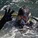 A diver is partially surfaced in open water, wearing full black tactical diving gear including a full-face mask and helmet-mounted light. The diver is making a hand signal with three fingers raised, likely communicating status or a number to someone off-camera. The image is sharp and well-lit, capturing fine details of the equipment and water surface.