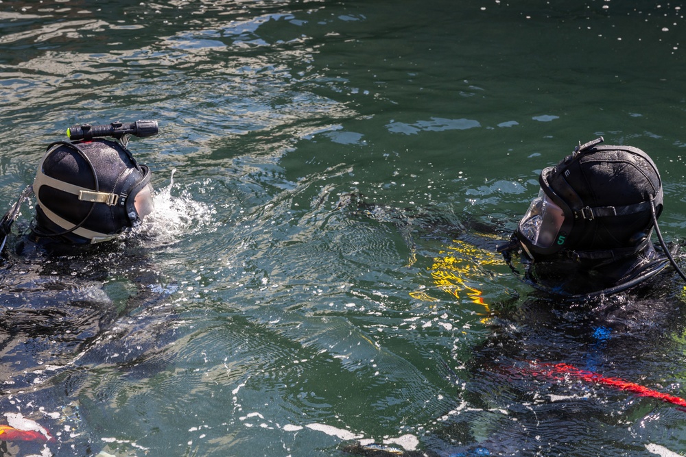 Two scuba divers are visible at the water surface in what appears to be a training or operational dive scenario. Both divers are wearing full black diving helmets with face masks and are equipped with diving gear. Colored ropes or lines (red, blue, yellow) are visible in the water near the right diver.