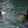 Forged Underwater: PNSY Dive Team Conducts Inspection of New Caisson Two scuba divers are visible at the water surface in what appears to be a training or operational dive scenario. Both divers are wearing full black diving helmets with face masks and are equipped with diving gear. Colored ropes or lines (red, blue, yellow) are visible in the water near the right diver.