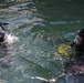 Two scuba divers are visible at the water surface in what appears to be a training or operational dive scenario. Both divers are wearing full black diving helmets with face masks and are equipped with diving gear. Colored ropes or lines (red, blue, yellow) are visible in the water near the right diver.