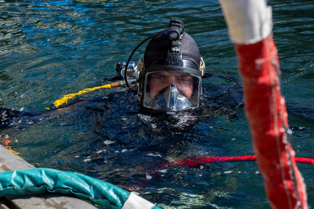 A diver is visible at the water surface wearing full professional diving equipment including a hard hat diving helmet with a clear face mask and mounted light. The diver appears to be in a harbor or controlled water environment, surrounded by various colored hoses and lines. The image captures a close-up perspective of the diver's face through the helmet visor.