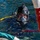 Forged Underwater: PNSY Dive Team Conducts Inspection of New Caisson A diver is visible at the water surface wearing full professional diving equipment including a hard hat diving helmet with a clear face mask and mounted light. The diver appears to be in a harbor or controlled water environment, surrounded by various colored hoses and lines. The image captures a close-up perspective of the diver's face through the helmet visor.