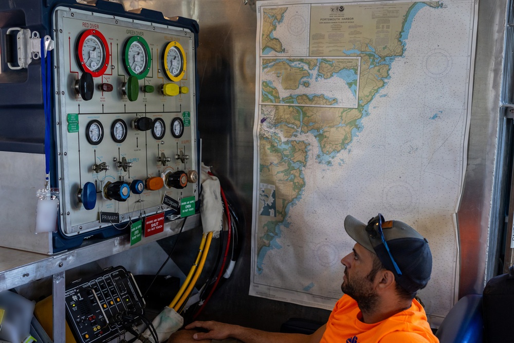 A man in an orange safety shirt and gray cap monitors a diving control panel inside what appears to be a dive support vessel or operations center. The panel features color-coded pressure gauges labeled 'RED DIVER' and 'GREEN DIVER', along with valves and air supply controls. A nautical chart of Portsmouth Harbor is mounted on the wall beside the control panel.