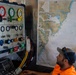 A man in an orange safety shirt and gray cap monitors a diving control panel inside what appears to be a dive support vessel or operations center. The panel features color-coded pressure gauges labeled 'RED DIVER' and 'GREEN DIVER', along with valves and air supply controls. A nautical chart of Portsmouth Harbor is mounted on the wall beside the control panel.