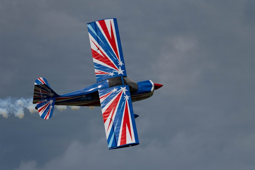 A small aerobatic aircraft is captured mid-flight in a banked or inverted position against an overcast sky. The plane features a bold patriotic paint scheme of red, white, and blue with star-burst designs. A white smoke trail is visible streaming from the tail, indicating an airshow performance.