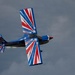 A small aerobatic aircraft is captured mid-flight in a banked or inverted position against an overcast sky. The plane features a bold patriotic paint scheme of red, white, and blue with star-burst designs. A white smoke trail is visible streaming from the tail, indicating an airshow performance.