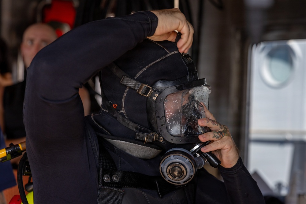 A person in a black wetsuit is donning or adjusting a full-face diving mask with an integrated regulator. The mask visor shows water droplets, suggesting recent use or wet conditions. The scene appears to take place aboard a vessel, with another person visible in the background.