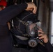 A person in a black wetsuit is donning or adjusting a full-face diving mask with an integrated regulator. The mask visor shows water droplets, suggesting recent use or wet conditions. The scene appears to take place aboard a vessel, with another person visible in the background.