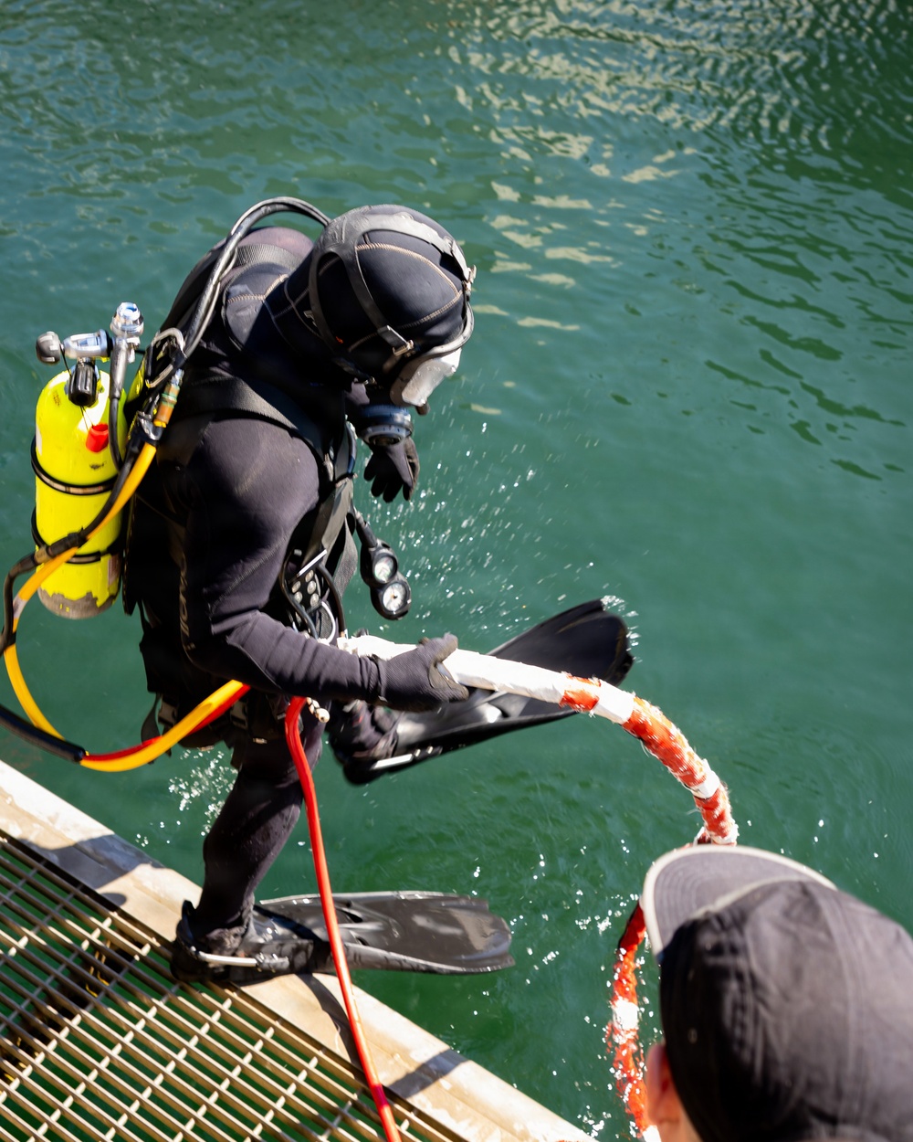 A fully equipped scuba diver in a black wetsuit is exiting or entering the water on a metal grating platform, holding a white and red braided rope. The diver is equipped with a bright yellow air tank, black fins, gloves, and a full-face diving helmet. A second person, visible only from the shoulders up wearing a gray cap, is assisting by holding the other end of the rope.