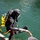 Forged Underwater: PNSY Dive Team Conducts Inspection of New Caisson A fully equipped scuba diver in a black wetsuit is exiting or entering the water on a metal grating platform, holding a white and red braided rope. The diver is equipped with a bright yellow air tank, black fins, gloves, and a full-face diving helmet. A second person, visible only from the shoulders up wearing a gray cap, is assisting by holding the other end of the rope.