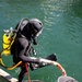 A fully equipped scuba diver in a black wetsuit is exiting or entering the water on a metal grating platform, holding a white and red braided rope. The diver is equipped with a bright yellow air tank, black fins, gloves, and a full-face diving helmet. A second person, visible only from the shoulders up wearing a gray cap, is assisting by holding the other end of the rope.