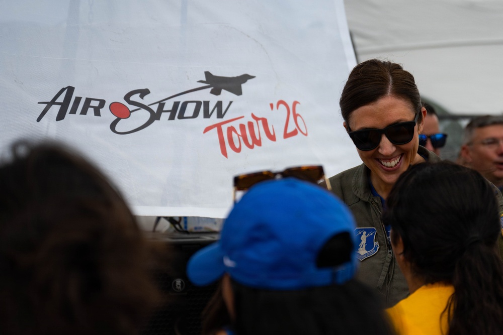 A smiling woman wearing sunglasses and an olive green military flight suit with a unit patch is interacting with a group of people, likely children, at an air show event. Behind her is a large white banner reading 'Air Show Tour '26' with a silhouette of a fighter jet. The scene appears to be a meet-and-greet or public engagement activity at an aviation event.
