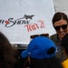 A smiling woman wearing sunglasses and an olive green military flight suit with a unit patch is interacting with a group of people, likely children, at an air show event. Behind her is a large white banner reading 'Air Show Tour '26' with a silhouette of a fighter jet. The scene appears to be a meet-and-greet or public engagement activity at an aviation event.