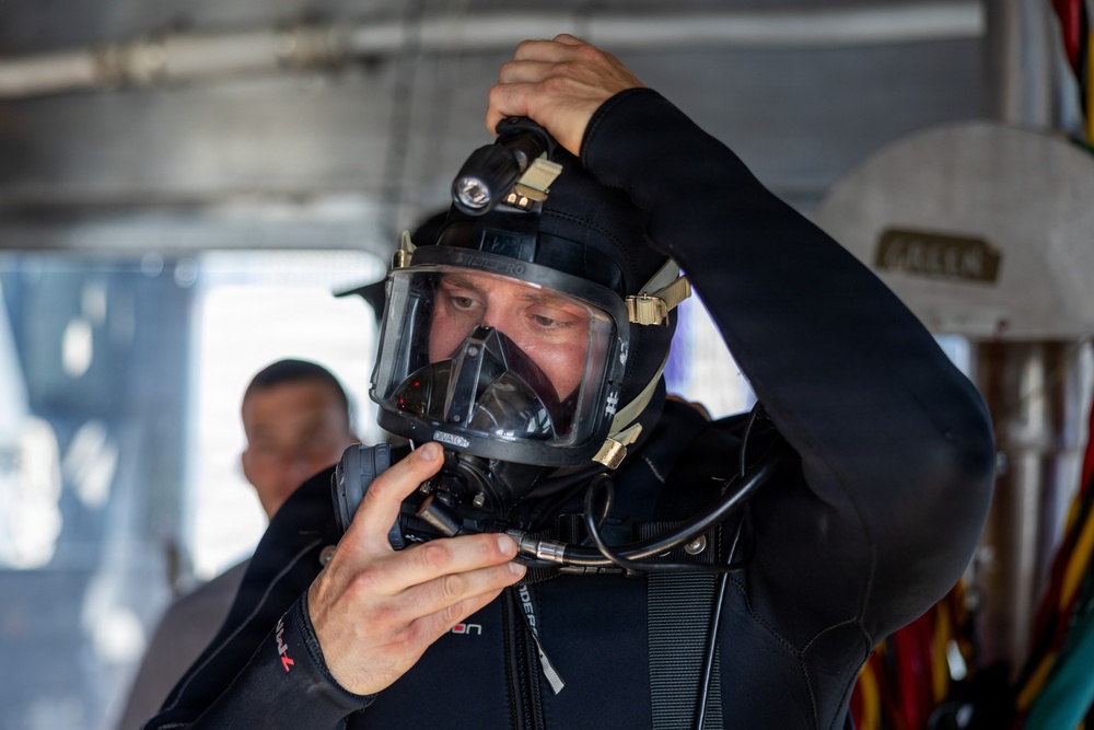 A diver in a black wetsuit is adjusting a full-face diving mask/helmet equipped with a mounted light and communication cables. The individual appears to be preparing for a dive, likely in a professional or military context given the advanced equipment. The setting appears to be aboard a vessel or covered platform with another person visible in the background.