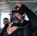 A diver in a black wetsuit is adjusting a full-face diving mask/helmet equipped with a mounted light and communication cables. The individual appears to be preparing for a dive, likely in a professional or military context given the advanced equipment. The setting appears to be aboard a vessel or covered platform with another person visible in the background.