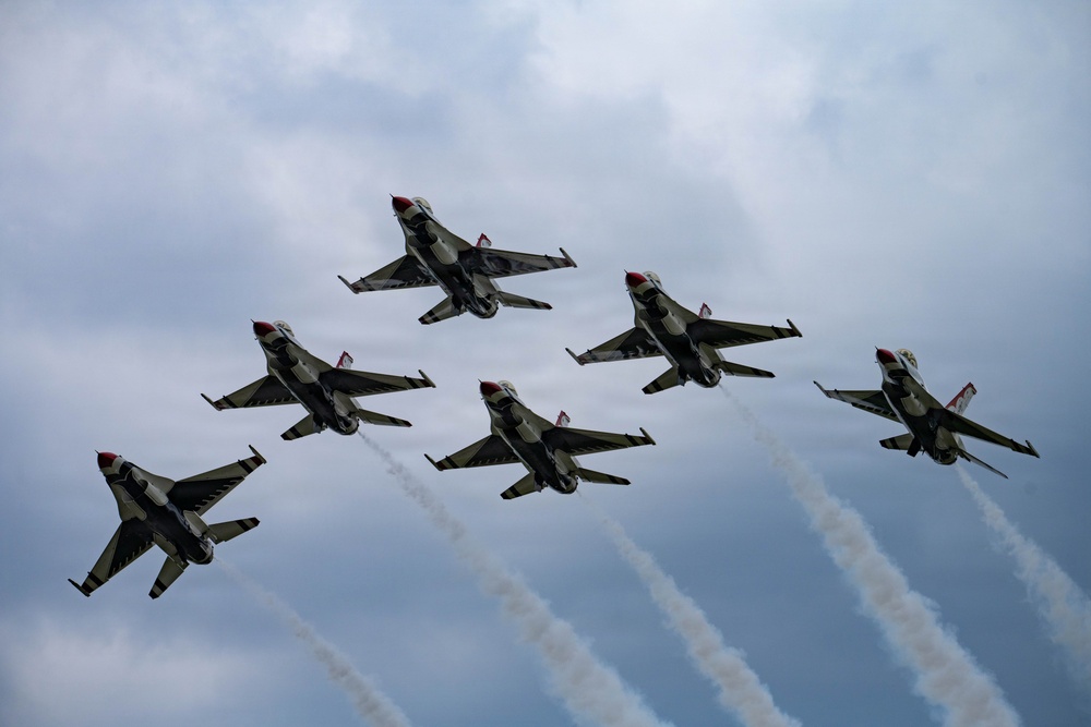 Six military jet aircraft are flying in a tight diamond/delta formation against an overcast sky. The aircraft display distinctive red, white, and blue livery consistent with the U.S. Air Force Thunderbirds aerial demonstration team. Multiple aircraft are emitting white smoke trails, indicating an active aerobatic performance.