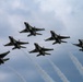 Six military jet aircraft are flying in a tight diamond/delta formation against an overcast sky. The aircraft display distinctive red, white, and blue livery consistent with the U.S. Air Force Thunderbirds aerial demonstration team. Multiple aircraft are emitting white smoke trails, indicating an active aerobatic performance.