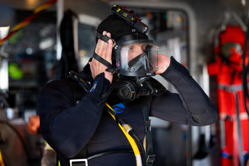 A diver in full black diving gear is adjusting a full-face diving mask while preparing for a dive. The individual is equipped with professional-grade equipment including a mounted headlamp, yellow air hose, and harness straps. The setting appears to be inside a vessel or dive support facility with red safety equipment visible in the background.
