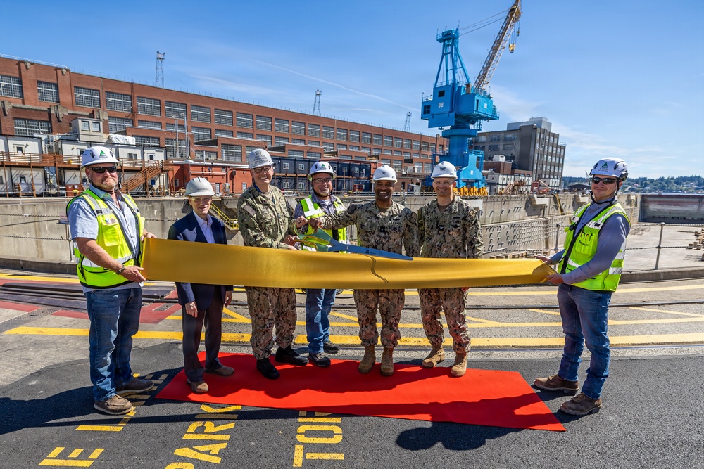 A ceremonial ribbon-cutting event is taking place at what appears to be a naval shipyard facility, with participants holding a large golden ribbon. The group consists of military personnel in Navy Working Uniform (NWU) camouflage, civilian contractors in high-visibility vests and hard hats, and at least one civilian official. A large blue crane and industrial brick buildings are visible in the background, consistent with a naval dry dock or shipyard environment.