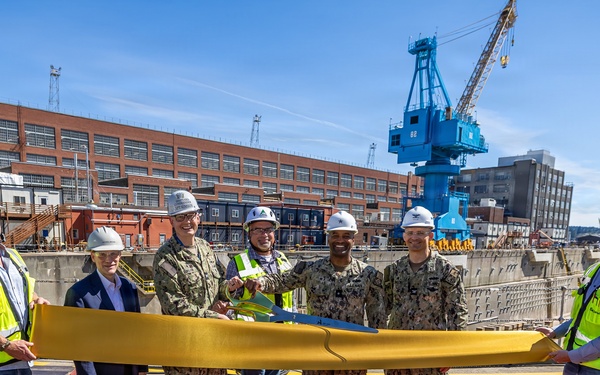 A ceremonial ribbon-cutting event is taking place at what appears to be a naval shipyard facility, with participants holding a large golden ribbon. The group consists of military personnel in Navy Working Uniform (NWU) camouflage, civilian contractors in high-visibility vests and hard hats, and at least one civilian official. A large blue crane and industrial brick buildings are visible in the background, consistent with a naval dry dock or shipyard environment.