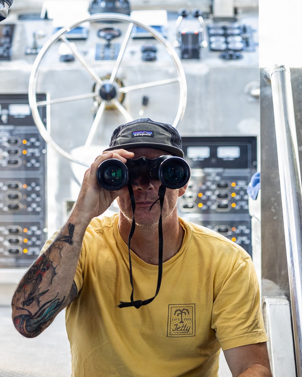 A tattooed man wearing a Patagonia cap and a yellow 'Jetty' branded t-shirt is looking through binoculars aboard what appears to be a vessel's bridge or helm station. Behind him is a large stainless steel ship's wheel and an extensive panel of navigation and control equipment. The image is sharp and well-lit, capturing a nautical observation scene.