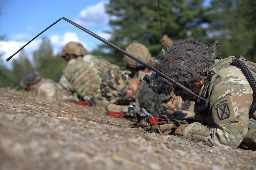 A group of soldiers in camouflage uniforms are lying prone on the ground in a firing line position. The foreground soldier clearly displays a '10th Mountain Division' patch on their right shoulder, and is equipped with a radio antenna and weapon. The scene appears to be a military training exercise conducted in an outdoor wooded environment.