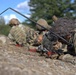 A group of soldiers in camouflage uniforms are lying prone on the ground in a firing line position. The foreground soldier clearly displays a '10th Mountain Division' patch on their right shoulder, and is equipped with a radio antenna and weapon. The scene appears to be a military training exercise conducted in an outdoor wooded environment.