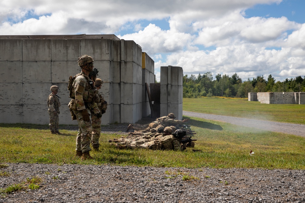 Military personnel in camouflage uniforms are conducting a live-fire training exercise at an outdoor range facility. Several soldiers are in the prone position firing weapons downrange while at least two others stand observing, likely in a supervisory or safety officer role. The setting includes concrete block structures typical of a military training installation.