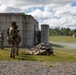 Military personnel in camouflage uniforms are conducting a live-fire training exercise at an outdoor range facility. Several soldiers are in the prone position firing weapons downrange while at least two others stand observing, likely in a supervisory or safety officer role. The setting includes concrete block structures typical of a military training installation.