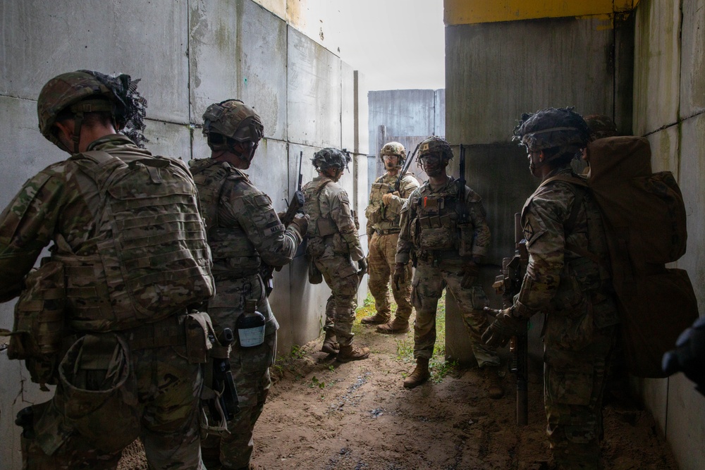 A group of approximately six military personnel in full combat gear are gathered in a narrow concrete corridor or alleyway between large concrete walls. They appear to be conducting a tactical briefing or coordination pause during what looks like a training exercise or operational movement. The soldiers are equipped with multicam uniforms, helmets, body armor, and weapons.