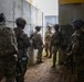 A group of approximately six military personnel in full combat gear are gathered in a narrow concrete corridor or alleyway between large concrete walls. They appear to be conducting a tactical briefing or coordination pause during what looks like a training exercise or operational movement. The soldiers are equipped with multicam uniforms, helmets, body armor, and weapons.