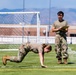 Two military personnel in OCP (Operational Camouflage Pattern) uniforms are on an athletic field, with one individual performing what appears to be a physical fitness test exercise near an orange cone while in a push-up or crawl position. A second individual stands upright holding a clipboard and pen, appearing to score or record the performance. The setting is an outdoor sports field with a soccer goal, football goal posts, and mountains visible in the background.