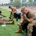 A group of military personnel in camouflage uniforms are resting on an outdoor artificial turf training area. They appear to be on a break, with several individuals looking at phones or tablets. The setting includes fitness equipment branded 'Rogue' and what appears to be a sports facility with chain-link fencing in the background.