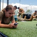 A group of military personnel in camouflage uniforms are taking a rest break on an outdoor artificial turf fitness area. The foreground shows a female soldier lying prone on the turf, focused on her smartphone, while several other soldiers sit or rest in the background also using phones. The setting appears to be a military base outdoor training facility with fitness equipment visible.