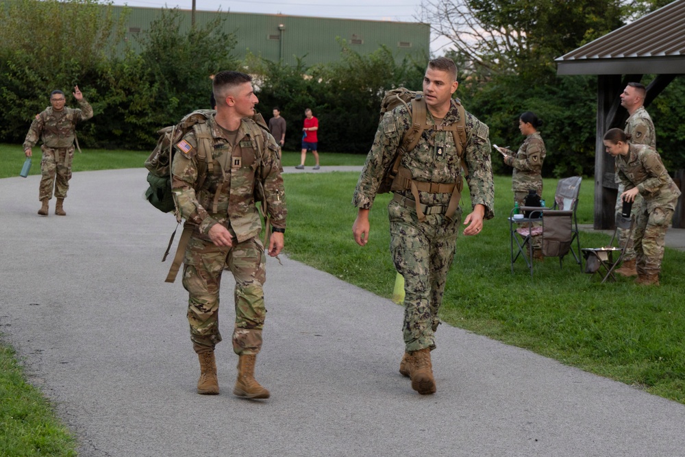 A group of military personnel in various camouflage uniforms are walking along a paved path in what appears to be a base or park setting. Two service members in the foreground — one in U.S. Army OCP (Operational Camouflage Pattern) and one in U.S. Navy NWU Type III (woodland digital) — are walking together while conversing and carrying rucksacks. Additional personnel are visible in the background near a covered pavilion area.