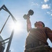 A low-angle upward shot captures a muscular individual pressing a hexagonal dumbbell overhead with one arm against a bright, sun-lit sky. The dramatic perspective emphasizes physical strength and determination. The setting appears to be an outdoor military or functional fitness training area.