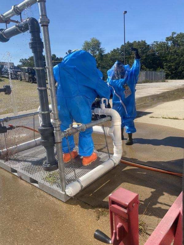 Two workers in blue hazmat/chemical protective suits and respirator masks are working on an industrial pipe system, with one leaning over the pipes and another standing nearby with one arm raised. Water or liquid is visibly spraying from the upper pipe connections, suggesting an active leak or pressurized system. The setting appears to be an outdoor industrial or municipal utility facility.